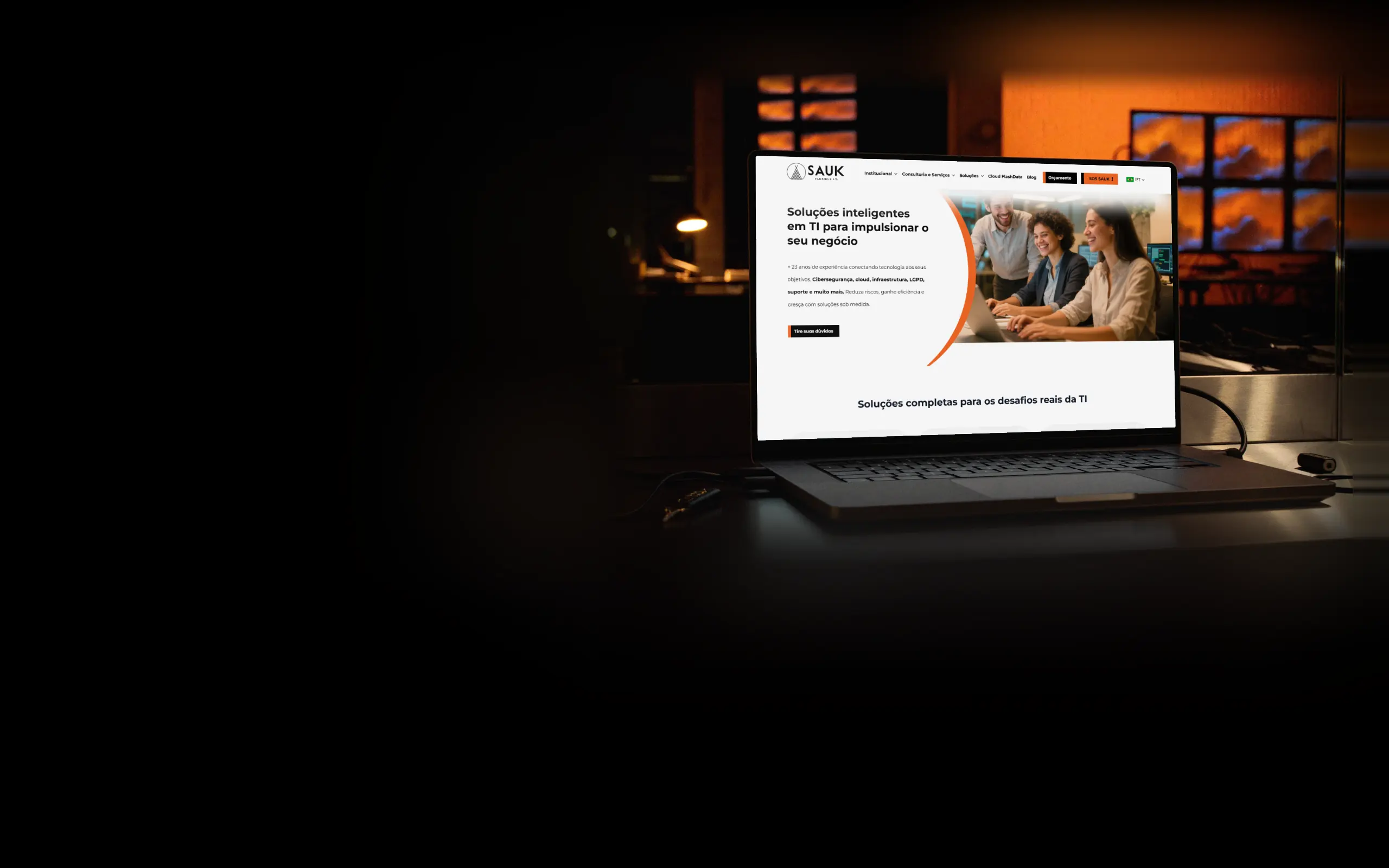 Notebook sobre a mesa de aço escovado de uma sala de monitoramento de TI à noite, com uma parede de telas em laranja e azul desfocada e uma chave de segurança USB ao lado, exibindo o site da Sauk